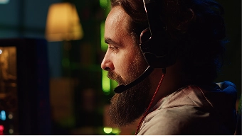 Een man met een baard en een koptelefoon zit in een slecht verlichte kamer, zijn blik strak op het computerscherm gericht. Het warme licht van een lamp en de groene achtergrond zorgen voor een intense gamesfeer.